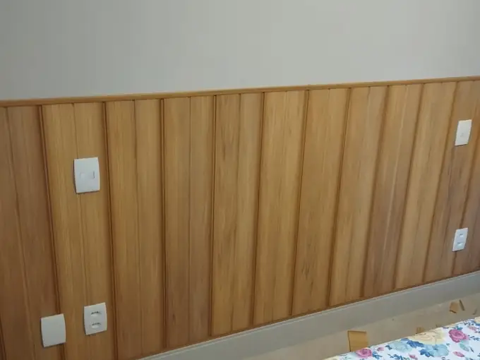 Revestimento de parede em PVC amadeirado em meia-parede de quarto, com recortes para interruptor e tomada